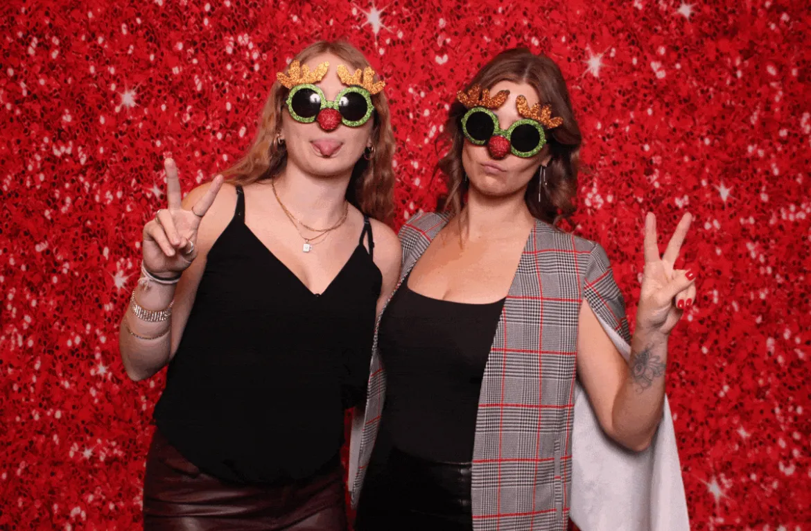 Two friends laughing together with reindeer-nose and antler prop glasses against a red sequin backdrop