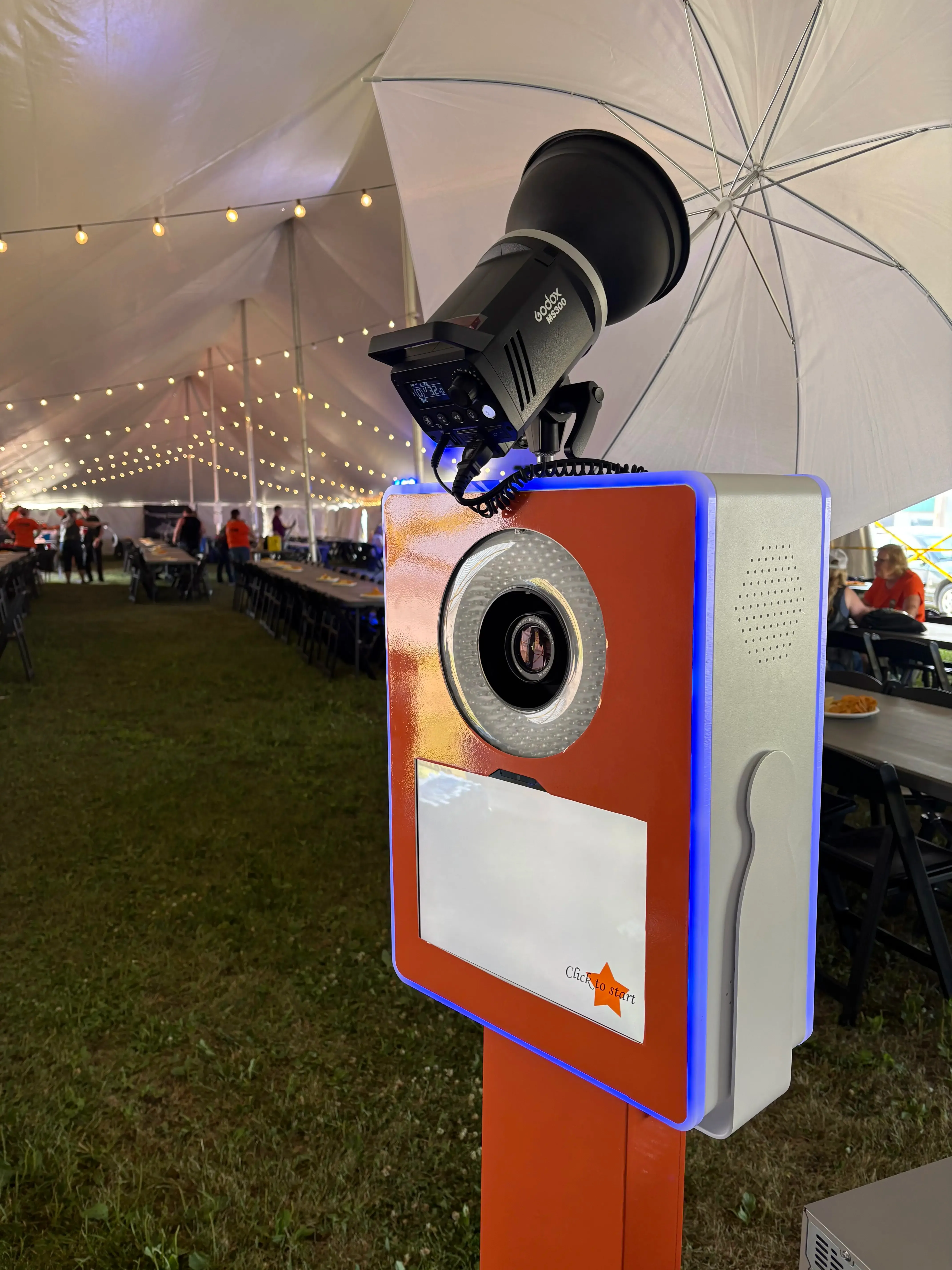 A self-serve photo booth kiosk with a touch display, set up for an outdoor corporate event under string-lit tents