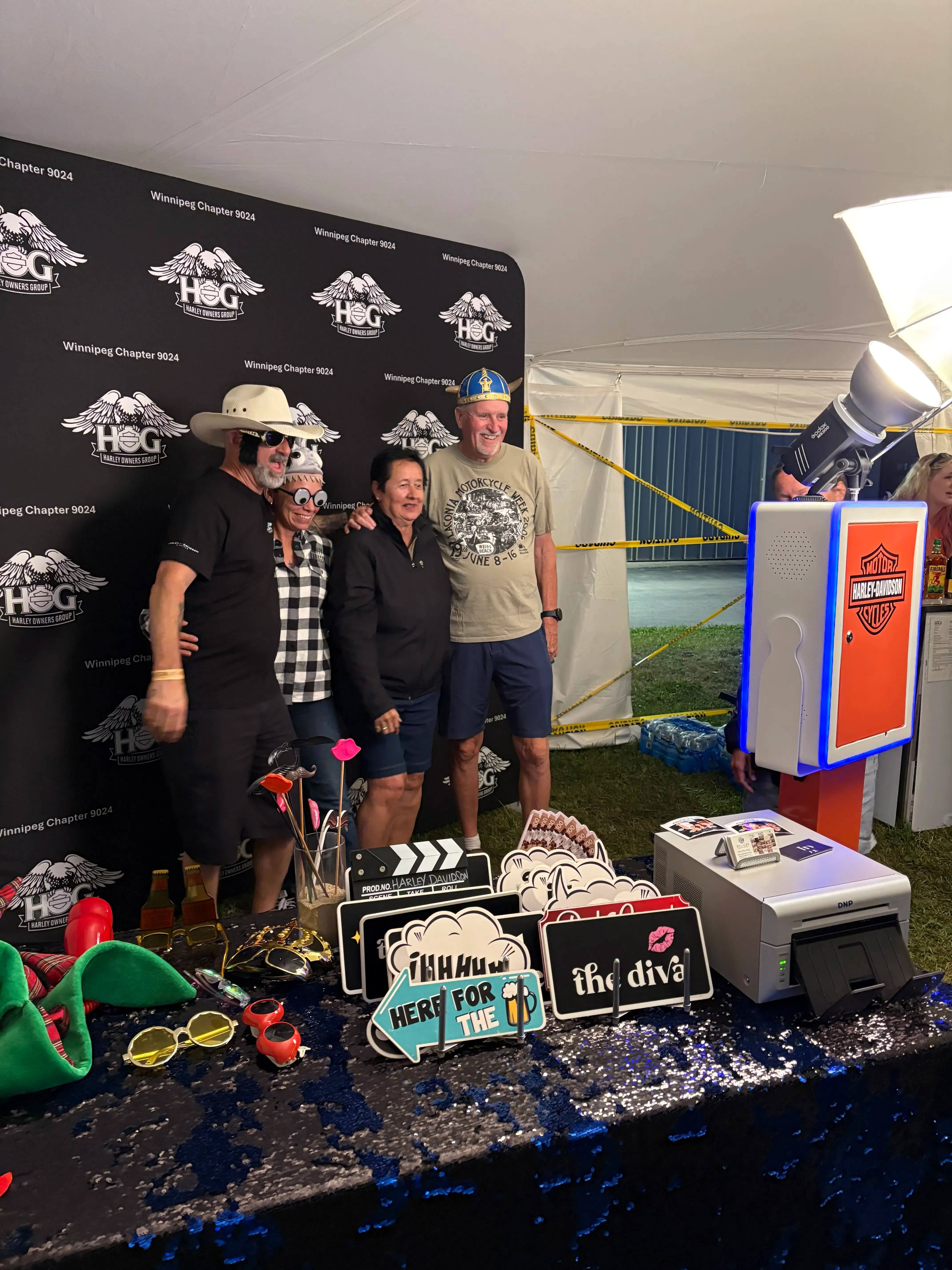 Guests striking fun poses with props in front of a custom Harley Owners Group, beside a branded photo booth and props table