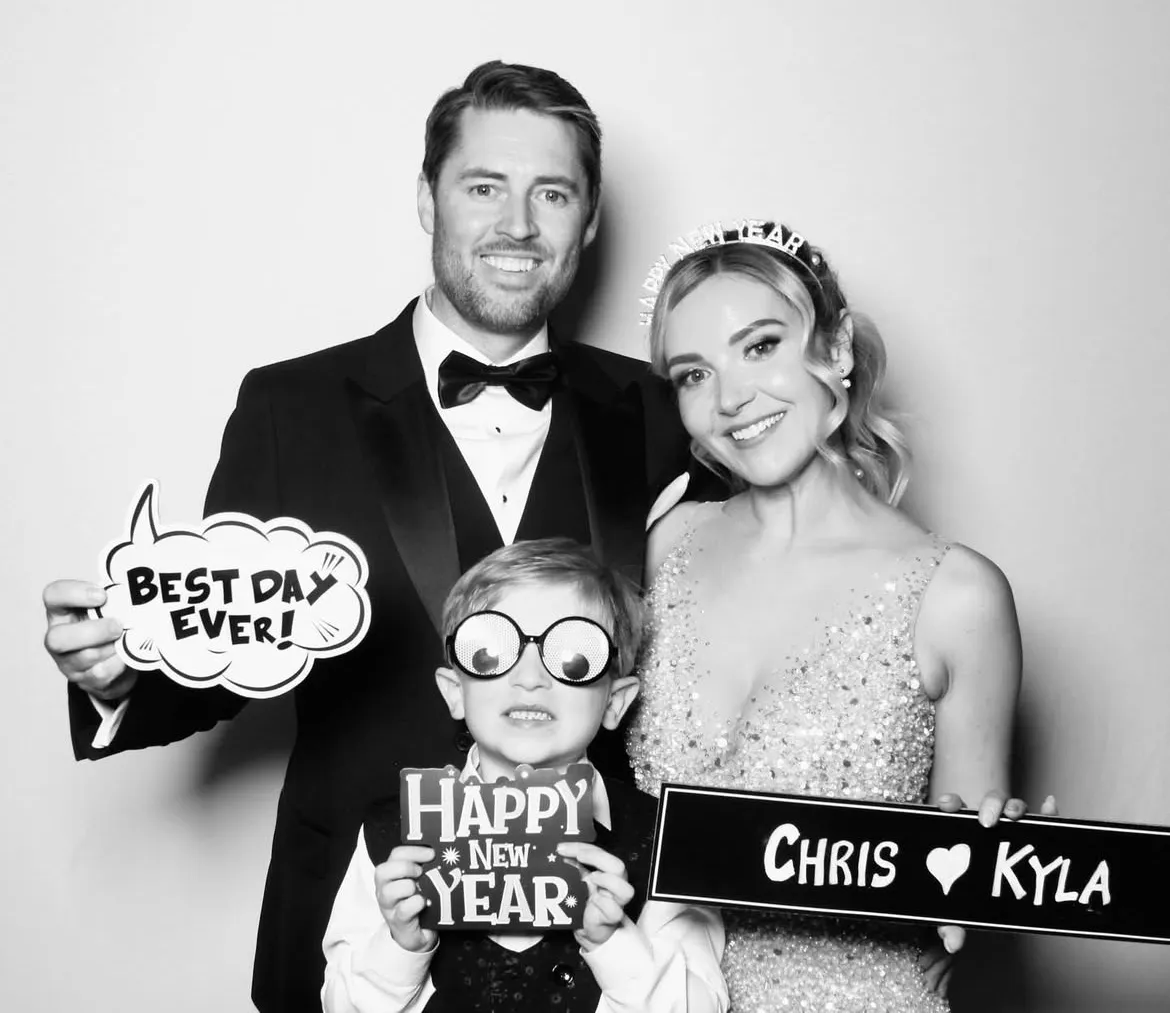 A tuxedoed guest and a young one posing with 'Best Day Ever' and 'Happy New Year' props