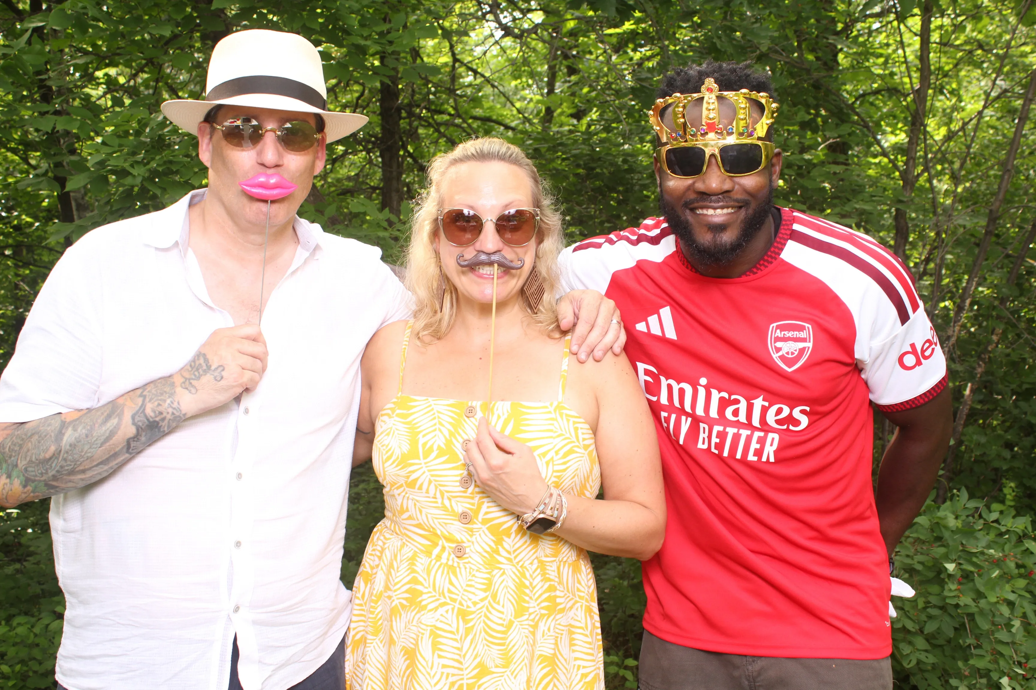 Three friends laughing with fun props at a sunny outdoor celebration surrounded by greenery