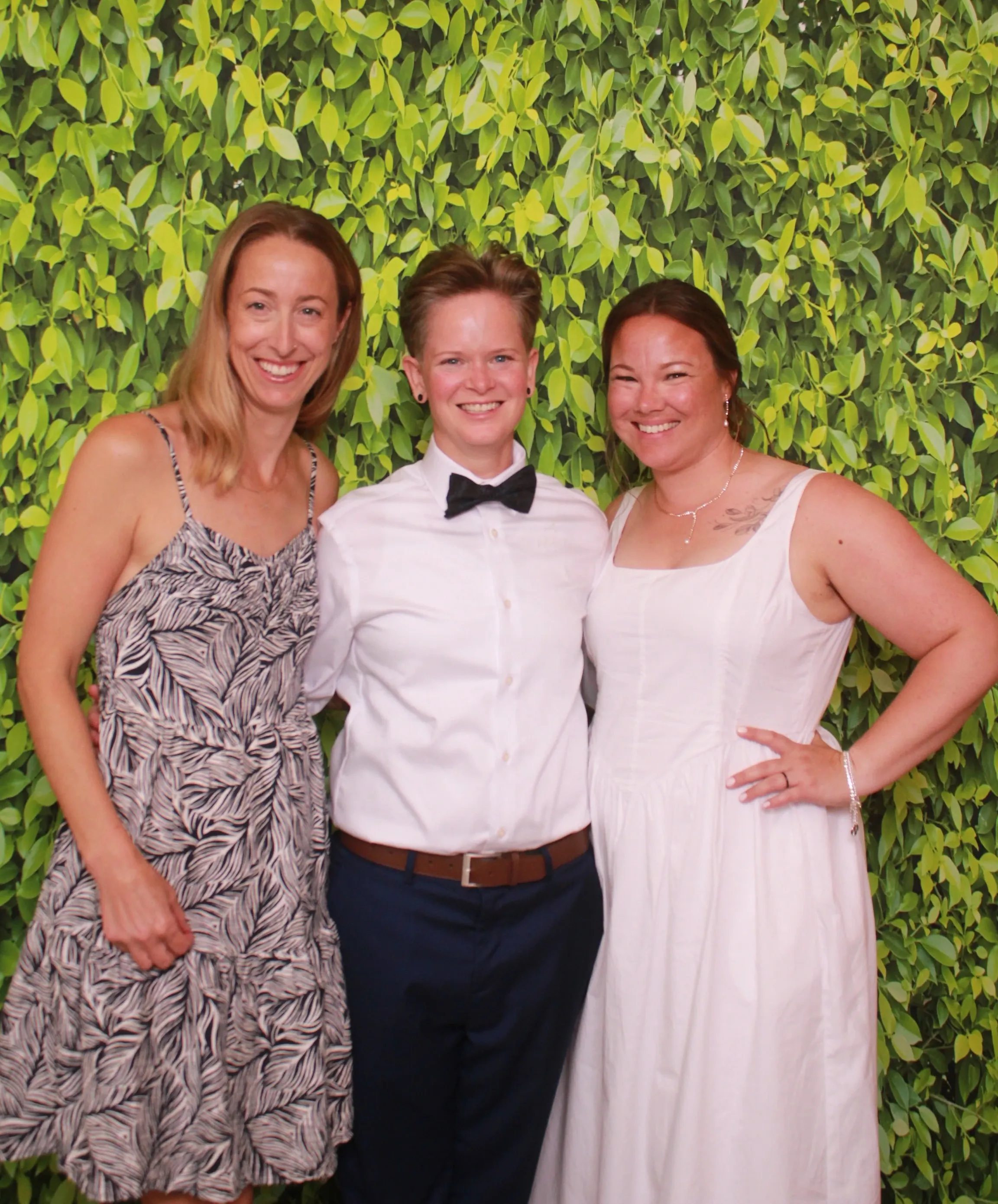 Three smiling friends posing in front of a lush green leafy backdrop