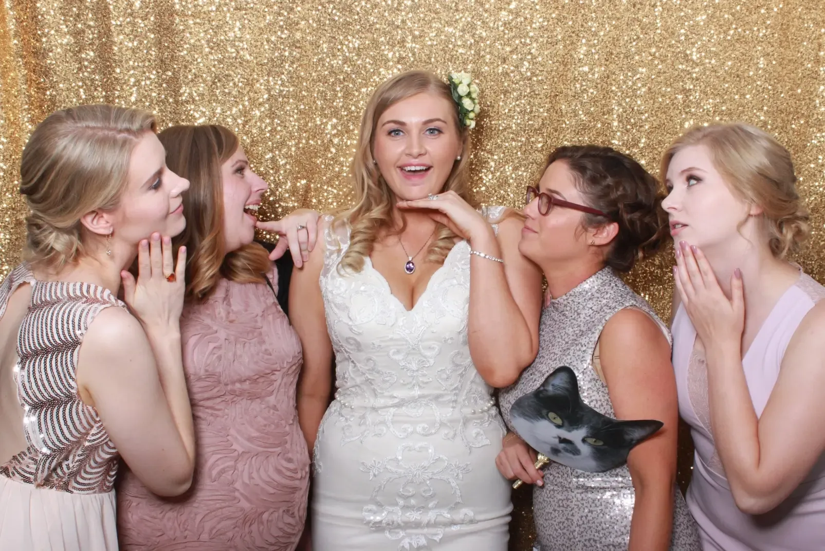 A bride surrounded by her bridesmaids, laughing with a cat-face prop in front of a gold sequin photo booth backdrop