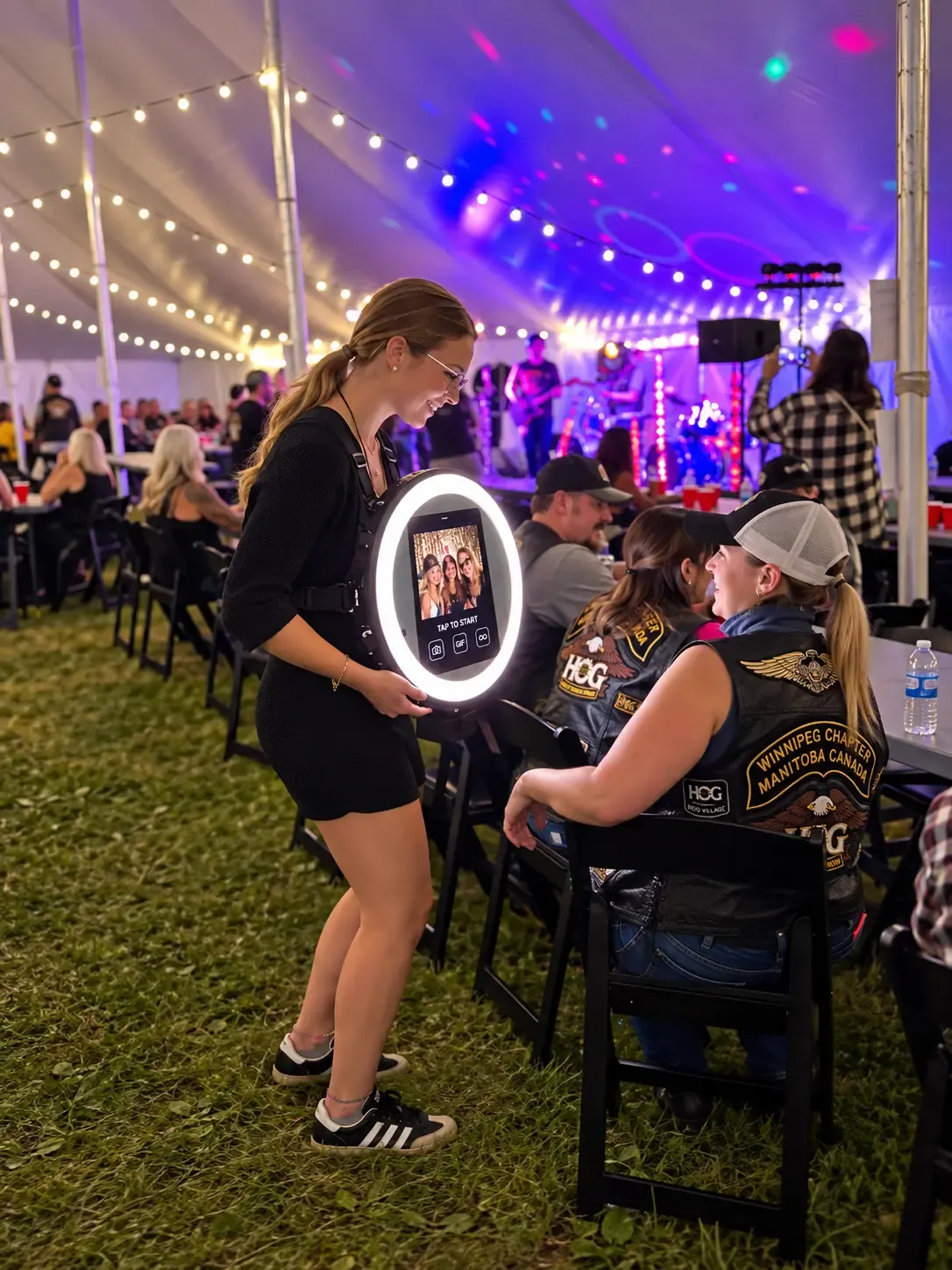 A roaming attendant photographing smiling guests with a handheld ring-light rig, live band and lights in the background